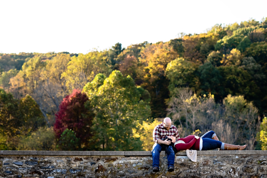 A pregnant couple lay on a stone wall with the mountains behind them.